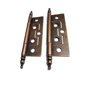 May include: Two antique brass hinges with decorative finials. The hinges are closed and have a distressed finish.