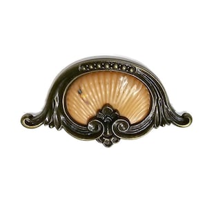 May include: Antique brass-finished drawer pull with an oval, shell-like center. The center is a light tan color with a fan-like design. The pull has ornate, curved edges and a decorative flourish at the bottom. The hardware is designed for furniture.
