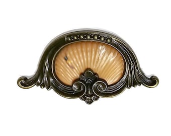 May include: Antique brass-finished drawer pull with an oval, shell-like center. The center is a light tan color with a fan-like design. The pull has ornate, curved edges and a decorative flourish at the bottom. The hardware is designed for furniture.