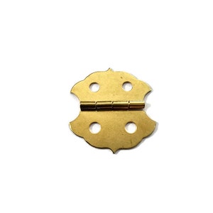 May include: A gold-toned metal hinge with a decorative, scalloped design. The hinge has four holes on each side and is closed.