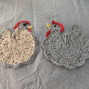 May include: Two crocheted chicken pot holders. One is grey and the other is beige with brown accents. Both have red combs and yellow beaks.