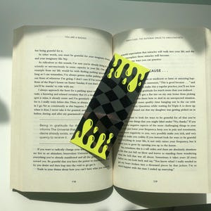 May include: A yellow and black bookmark with a dripping paint design. The bookmark is placed on an open book with the text "You are a badass" and "Gratitude: The Gateway Drug to Awakeness".