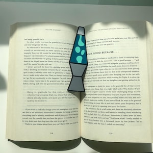 May include: A blue and green lava lamp bookmark with the text "This is good because..." on the right side. The bookmark is placed between the pages of an open book.