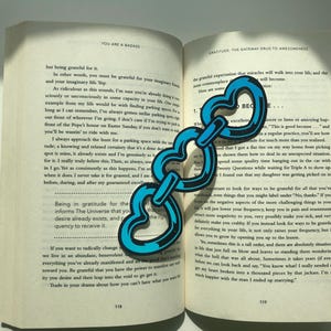 May include: A blue heart-shaped bookmark with a chain design. The bookmark is placed on an open book with text about gratitude and being grateful.