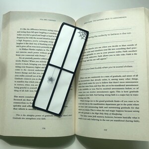 May include: A black and white bookmark featuring a spider web design. The bookmark is made from clear material with a black frame. The bookmark is positioned on an open book with the text "Gratitude: The Gateway Drug to Awesomeness" visible.