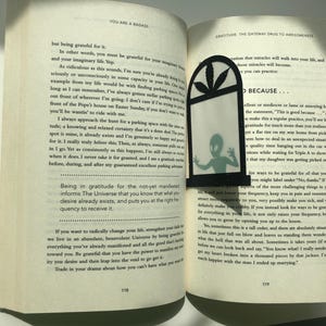 May include: An open book with a black and green bookmark featuring a silhouette of an alien. The book's pages contain text about gratitude and the universe. The text on the bookmark reads "Because..."