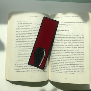 May include: A red and black bookmark with a starburst design. The bookmark is placed on an open book with the text "GOOD BECAUSE..." visible on the page.