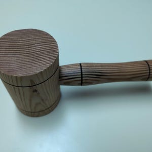 May include: A wooden mallet with a round head and a long handle. The head is made of light brown wood with dark brown stripes. The handle is made of the same wood and has two black lines around it.