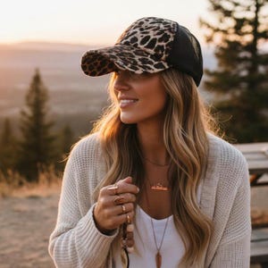 May include: A baseball cap with a leopard print design and a black mesh back. The person is wearing a cream-coloured cardigan and a white top. The person is also wearing several rings and necklaces.