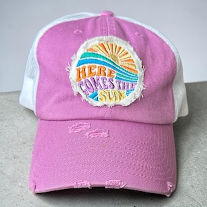 May include: A pink baseball cap with a white mesh back. The front of the cap features a distressed patch with the text "HERE COMES THE SUN" and a colourful sun and wave design. The cap has a distressed brim.