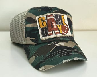 May include: A camouflage print trucker hat with a mesh back and a distressed brim. A rectangular patch on the front features the text GAME DAY in yellow and dark brown, with a brown football graphic replacing the letter A and a football icon.