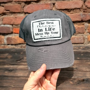 May include: A charcoal grey distressed baseball cap with a mesh back and a rectangular white patch on the front. The patch features black text that reads, The Best Things In Life Mess Up Your Hair, with frayed edges and visible stitching.