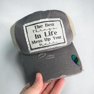 Pode incluir: Um boné de beisebol cinza com parte traseira de malha bege e aba desgastada. Um patch retangular branco está costurado na frente com o texto preto: The Best Things In Life Mess Up Your Hair. Um pequeno adesivo holográfico está na aba.
