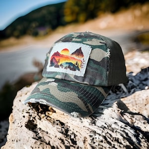 Puede incluir: Una gorra de camionero de camuflaje con un aspecto desgastado. La gorra tiene un parche con una escena de montaña colorida y una puesta de sol.