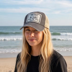 May include: A charcoal gray distressed baseball cap with a mesh back and a rectangular white patch on the front. The patch features black text that reads, The Best Things In Life Mess Up Your Hair, with a frayed edge design on the fabric.