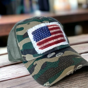 May include: Camouflage baseball cap with a distressed design. Features a patch of the American flag with a frayed edge. The cap has a mesh back and a curved brim, suitable for outdoor activities.