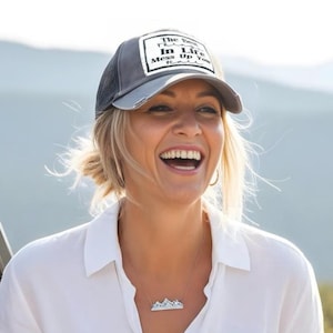 Peut inclure: Une casquette de type trucker gris anthracite avec un écusson rectangulaire blanc portant le texte noir The Best Things In Life Mess Up Your Hair. La casquette possède un dos en filet et une visière courbée. Un collier pendentif montagne est visible.