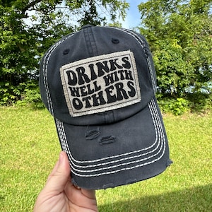 May include: A distressed navy blue baseball cap with white stitching and a frayed brim. The cap features a rectangular patch with the words "DRINKS WELL WITH OTHERS" in a retro font. The hat is worn and has a vintage look.