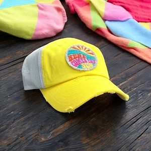 May include: Yellow trucker hat with a white mesh back and a colorful embroidered patch that says "Here Comes The Sun".