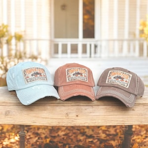 Puede incluir: Tres gorras de béisbol desgastadas en azul claro, óxido y marrón, cada una con un parche de "Pumpkin Patch". El parche presenta un camión, calabazas y texto. Las gorras están sobre una superficie de madera.