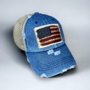 Puede incluir: Una gorra de béisbol de mezclilla azul con un panel trasero de malla y un parche de bandera estadounidense desgastado. La gorra tiene una visera curva y un borde deshilachado.