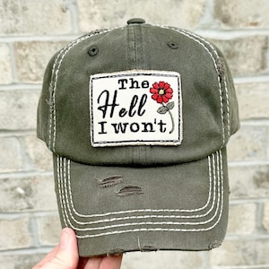 May include: An olive green baseball cap with a distressed finish and white contrast stitching. A rectangular patch on the front features the embroidered black text "The Hell I won't" alongside a small red flower with a green stem and leaves.