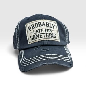 May include: Navy blue distressed baseball cap with white stitching and a pre-curved bill. The cap features a rectangular patch with the words "PROBABLY - LATE FOR - SOMETHING" in black text.