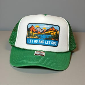 Op de afbeelding: Groene en witte truckerpet met een rechthoekige patch met een landschap met bergen, een meer en bomen. De patch heeft de tekst "LET GO AND LET GOD" in witte letters. De pet heeft een groene klep en een mesh achterkant.