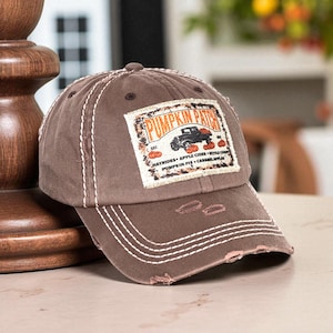 Peut inclure: Casquette de baseball marron délavée avec des coutures blanches et un écusson rectangulaire. L'écusson indique "Pumpkin Patch" avec un dessin de camion, de citrouilles et du texte. La casquette a une visière incurvée et un aspect vieilli.