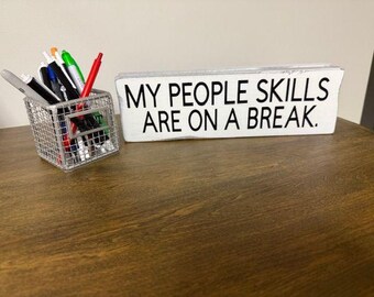 May include: A white wooden sign with the text "MY PEOPLE SKILLS ARE ON A BREAK" in black lettering. A small, silver wire basket filled with pens and markers sits to the left of the sign on a wooden desk.