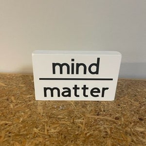 May include: White wooden sign with black text that reads "mind over matter".