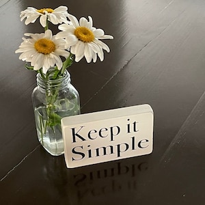 May include: A white sign with the text "Keep it Simple" on a dark wood surface with a small glass vase filled with white daisies.