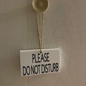 May include: White sign with black text that reads "PLEASE DO NOT DISTURB". The sign is hanging from a string attached to a small, round, cream-colored knob.