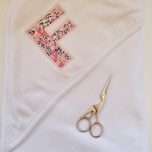 May include: A white hooded towel with a pink floral letter F embroidered on it. A pair of silver scissors are lying on the towel.
