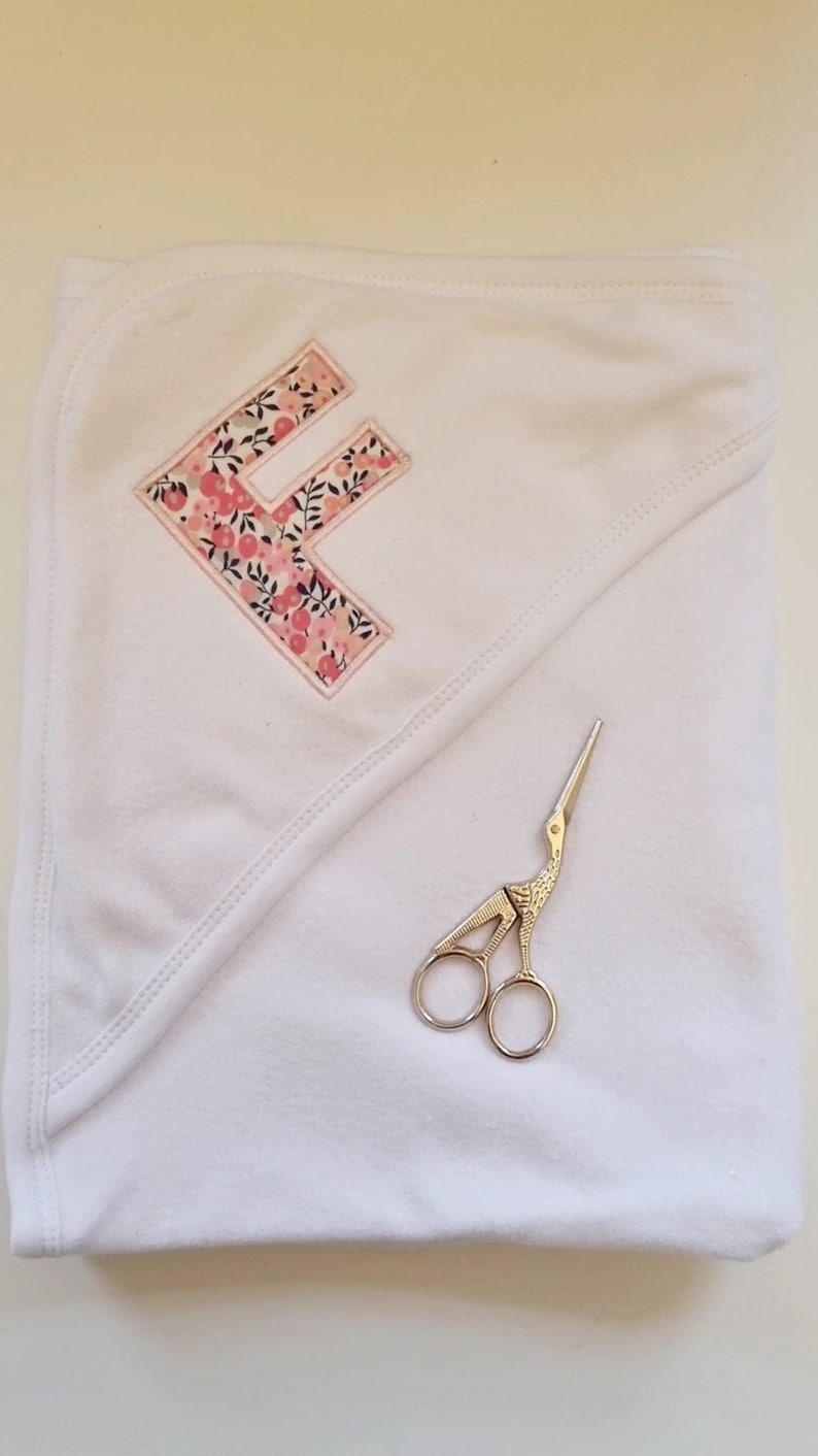 May include: A white hooded towel with a pink floral letter F embroidered on it. A pair of silver scissors are lying on the towel.