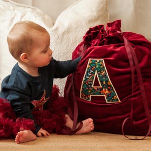 May include: A large, deep red velvet drawstring gift sack featuring a prominent, embroidered letter A on the front. The letter A is decorated with a colourful, intricate floral and botanical pattern in shades of green, blue, and orange.