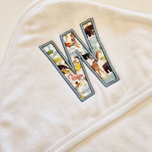 May include: White hooded towel with a blue outlined letter W embroidered on the front. The letter W is filled with a colorful pattern of animals.