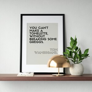 Może przedstawiać: Czarny obraz w ramie z białym tłem i czarnym tekstem, który brzmi "You can't make a tomette without breaking some Greggs. Tom Wambsgans." Obraz znajduje się na drewnianej półce z złotą lampą i rośliną w doniczce.