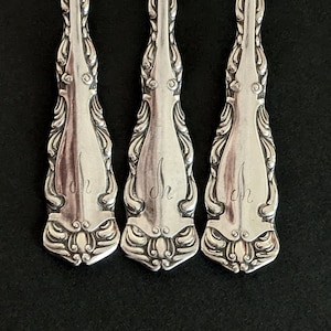 May include: Close-up of three silver spoons with ornate, floral-like designs on the handles. Each spoon has the initials "Jh" engraved on the handle. The spoons are arranged vertically against a dark background.
