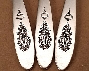 May include: Three silver spoons with ornate black engraved designs. Each spoon features a decorative emblem at the top and a detailed floral pattern. The spoons are arranged on a brown surface, showcasing their elegant design.
