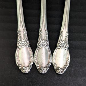 May include: Close-up of three silver spoons with ornate floral and scrollwork detailing. The spoons have an oval bowl shape and a decorative handle. The spoons are arranged vertically against a dark background.