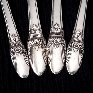 May include: Close-up of four silver spoons with ornate floral and geometric designs on the handles. The spoons are arranged diagonally against a dark background, showcasing the intricate details of the silverware.