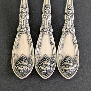 May include: Three antique silver forks with ornate handles and tines. The handles feature a detailed grape cluster design. The forks are arranged on a dark surface, showcasing their intricate craftsmanship and vintage appeal.