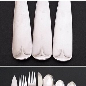 May include: Set of seven silver-toned flatware pieces, including a butter knife, two forks, a spoon, and three teaspoons. The flatware is arranged on a black surface.