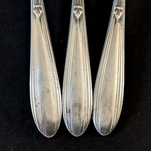 May include: Close-up of three silver spoons with a decorative design on the handle. The spoons have a classic, elegant shape with a slightly tarnished appearance, suggesting age or use. The background is a solid black, providing contrast to the silverware.