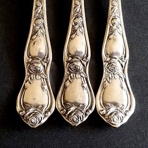 May include: Three silver forks with ornate rose designs on the handles. The forks are arranged in a row, with the handles pointing upwards.