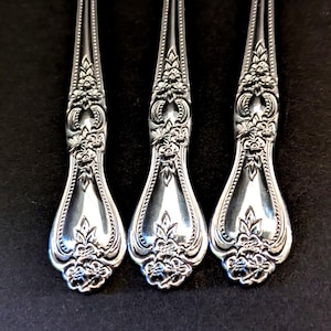 May include: Three ornate silver spoons with floral and scrollwork detailing. The spoons have a teardrop-shaped bowl and a decorative handle. The spoons are arranged on a dark surface, showcasing their intricate design.