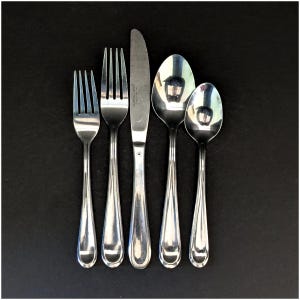 May include: A set of five stainless steel utensils: two forks, a knife, and two spoons. The silverware is arranged vertically against a dark background, reflecting light. The utensils have a simple, elegant design.