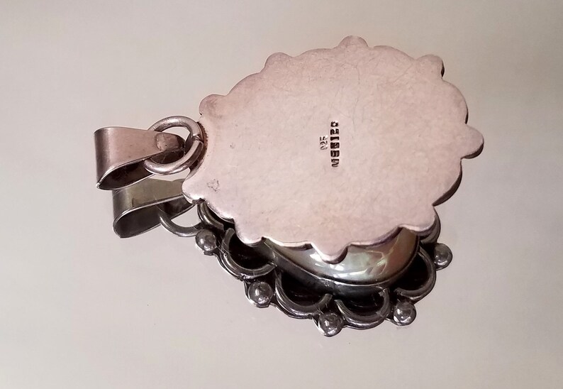 May include: Silver pendant with an ornate design and a silver cabochon stone. The back of the pendant is stamped with "925" and "MADE IN ITALY".