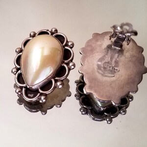 May include: A pair of silver earrings with a pearlescent white teardrop-shaped stone set in a silver filigree design. The earrings have a clip-on closure.